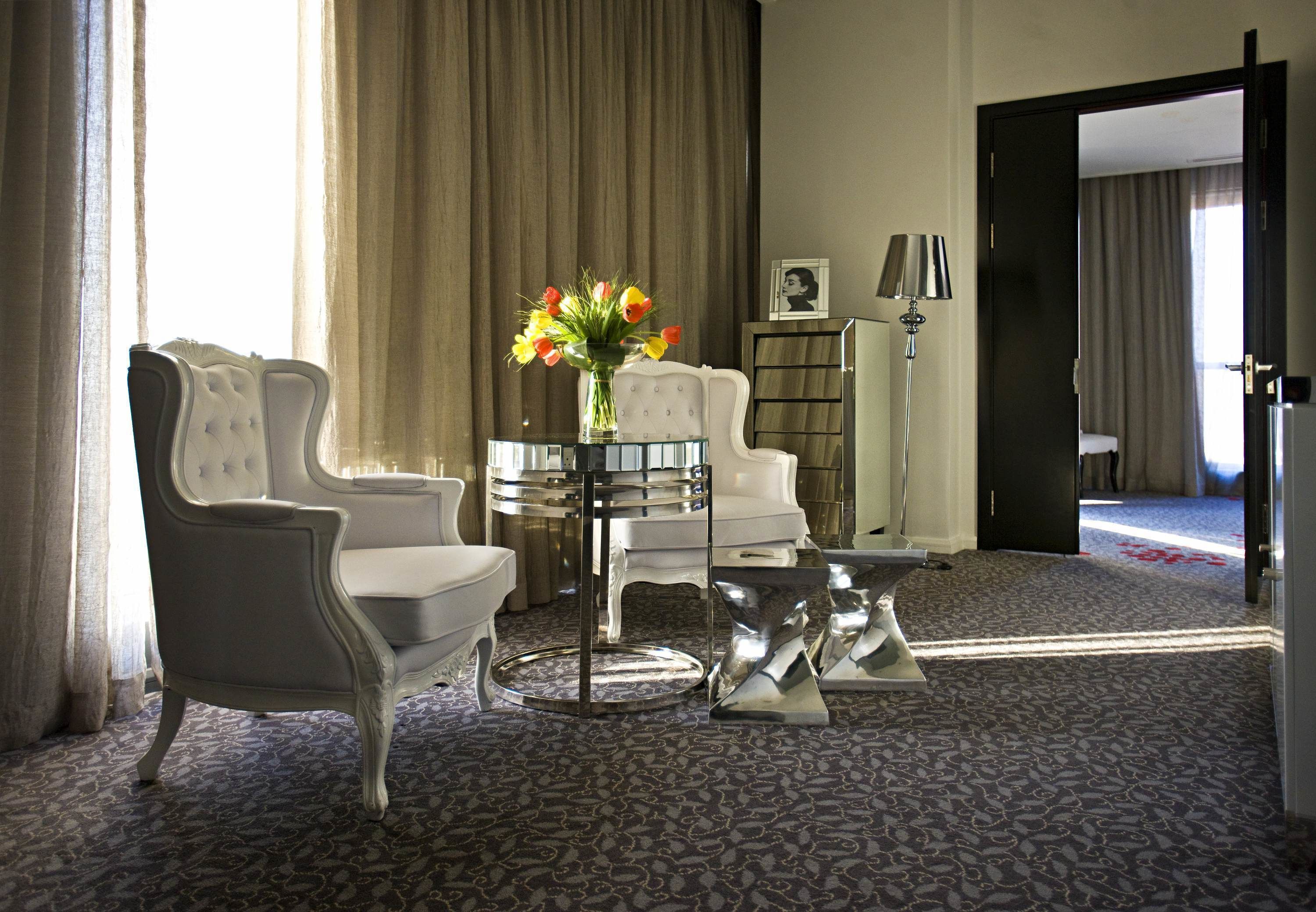 Ein Hotelzimmer mit großem Fenster, einem weißen Sessel, einem kleinen runden Tisch mit Blumenvase und einem Bücherregal.