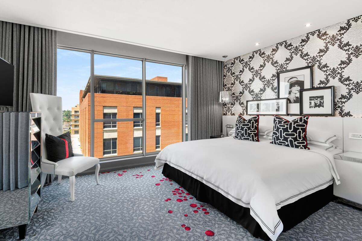 Une chambre d’hôtel moderne dotée d’un grand lit, d’un coin salon avec un fauteuil, d’une télévision et d’une grande fenêtre donnant sur un immeuble en briques.