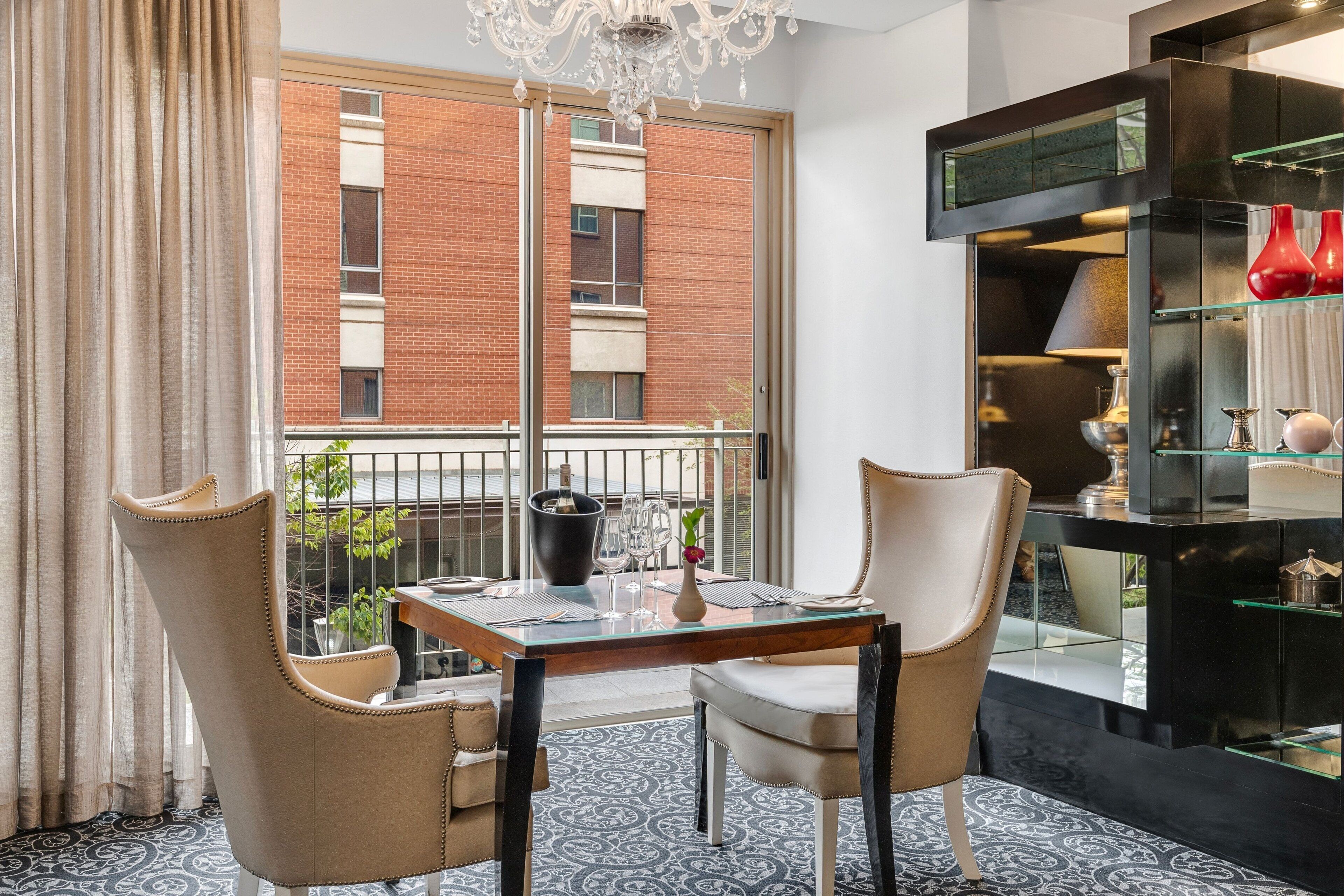 Un coin repas avec une table en verre, deux chaises, un lustre et une vue sur un bâtiment en briques.