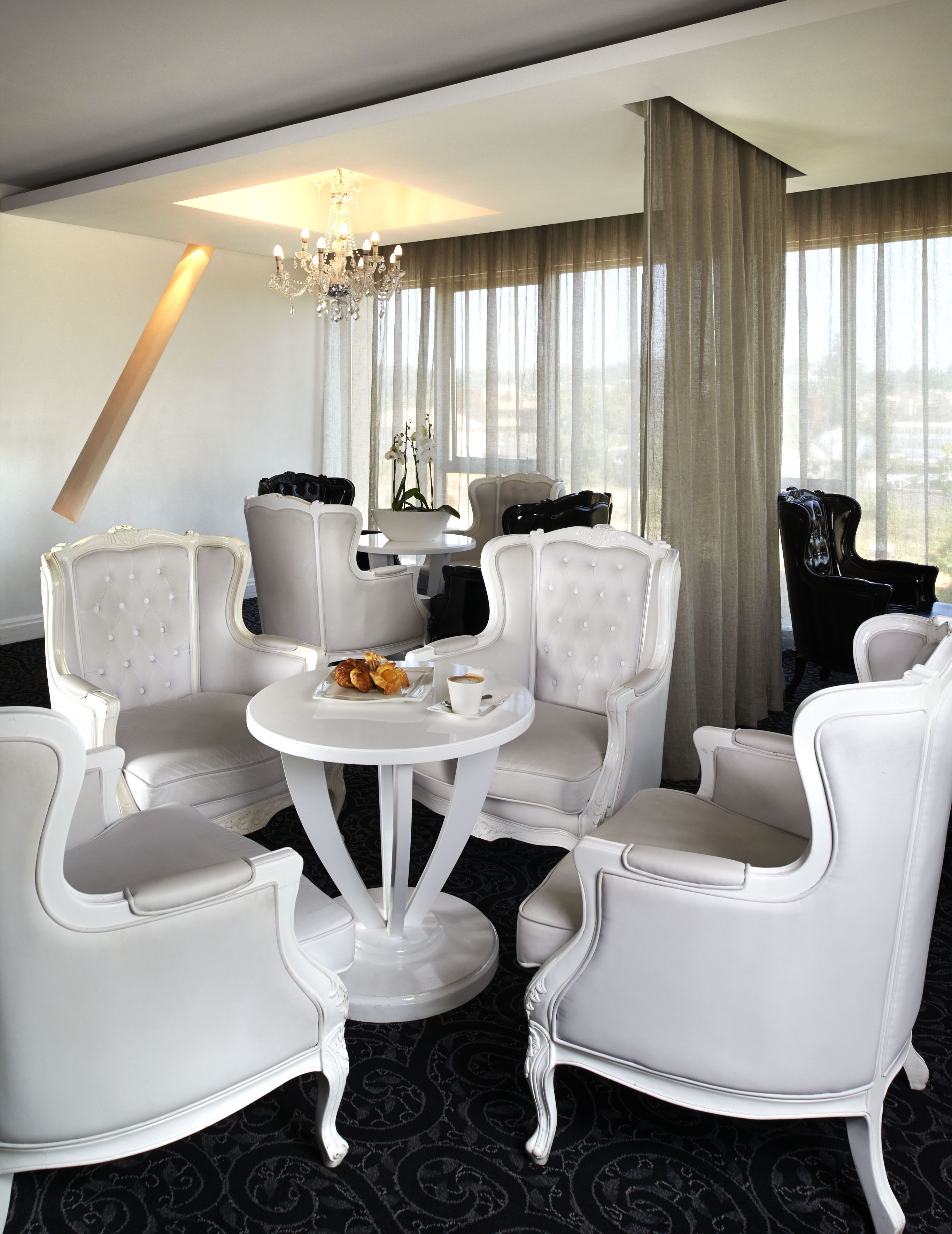 Una habitación de hotel con una mesa redonda blanca rodeada de cuatro sillas blancas tapizadas, una araña y vistas de la ciudad a través de amplias ventanas con cortinas de encaje.