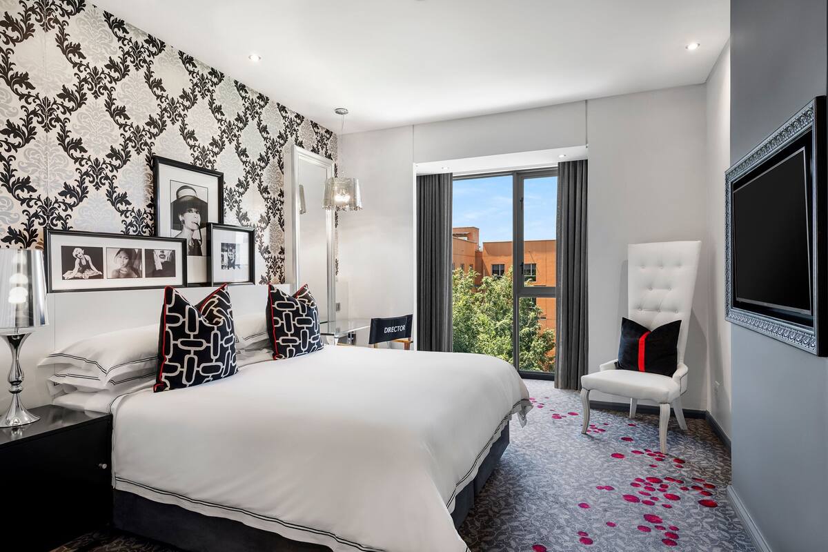 Une chambre d’hôtel moderne avec un grand lit, un fauteuil blanc, une télévision et une vue sur les immeubles par une grande fenêtre.