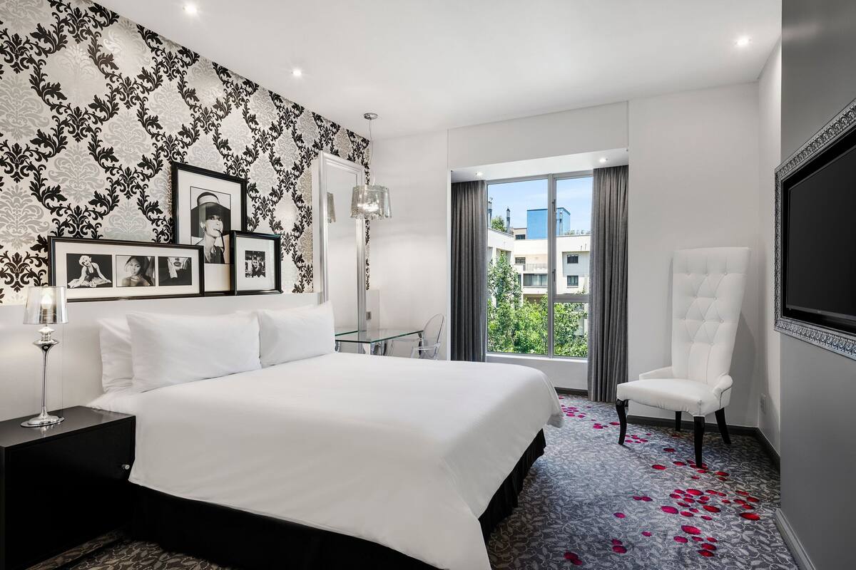 Une chambre d’hôtel moderne avec un grand lit, une table en verre, un fauteuil blanc et un téléviseur fixé au mur.