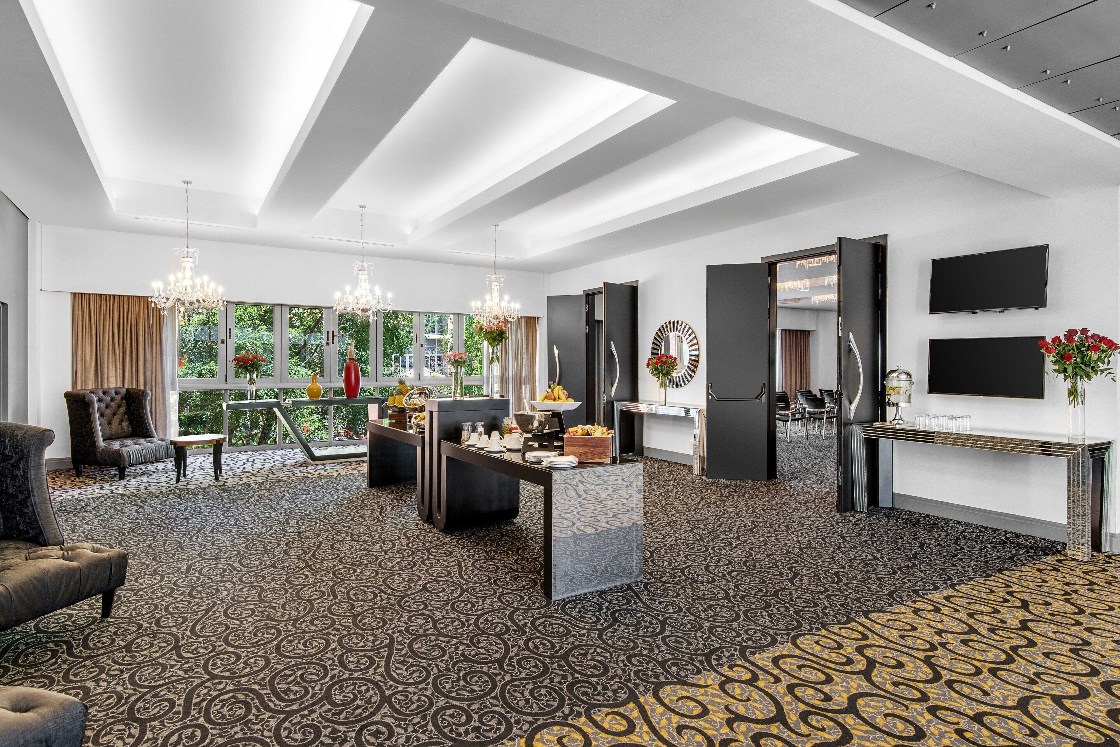 A spacious hotel lobby with a patterned carpet, a reception desk, a seating area with a sofa and armchairs, a chandelier, and a large window offering a view of greenery.
