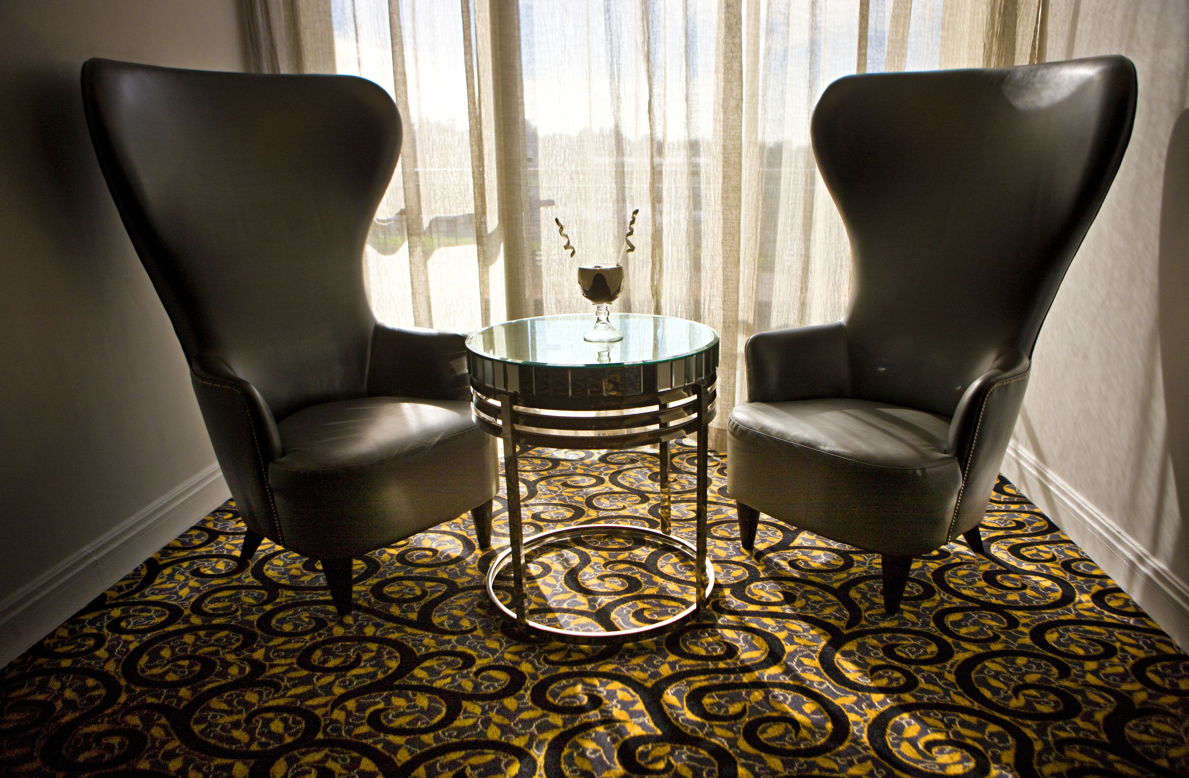 Two leather armchairs facing each other around a glass table with a decorative item on it, set on a patterned carpet in a room with a large window and sheer curtains.