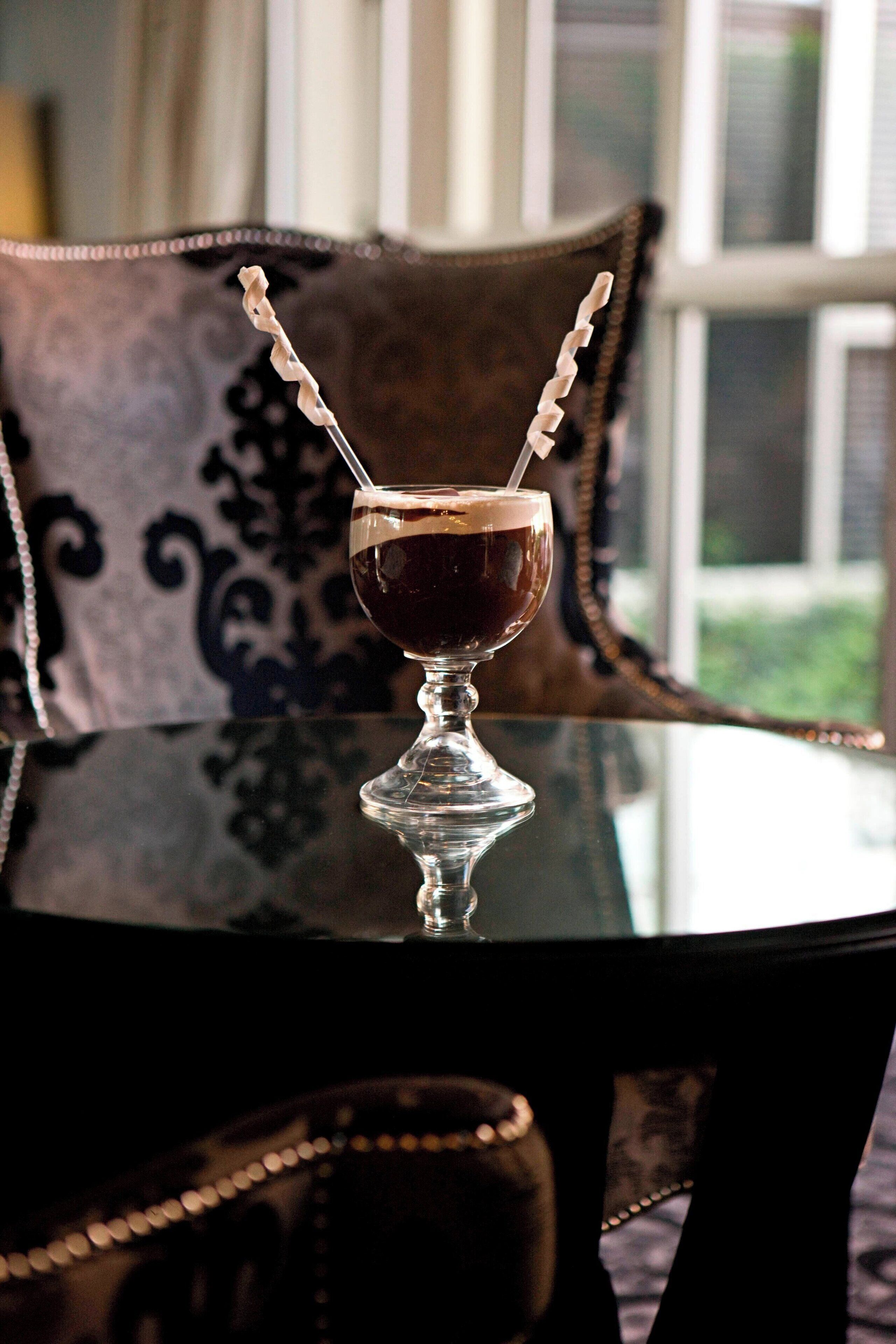 A glass with a cocktail on a reflective table, set against a patterned chair and a window with a view.