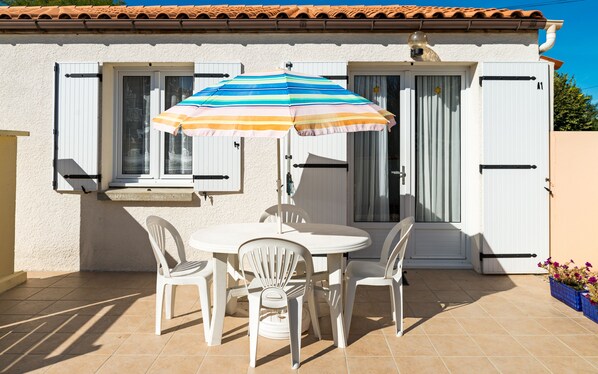 A patio with a white table and four white chairs, a striped umbrella, and a white building with shutters.
