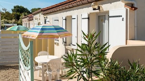 A row of white-framed rooms with a tiled roof, a small outdoor seating area with a table and chairs, and a striped umbrella providing shade. - Résidence Lagrange Vacances Les Maisons de Saint-Georges (Saint-Georges-de-Didonne)