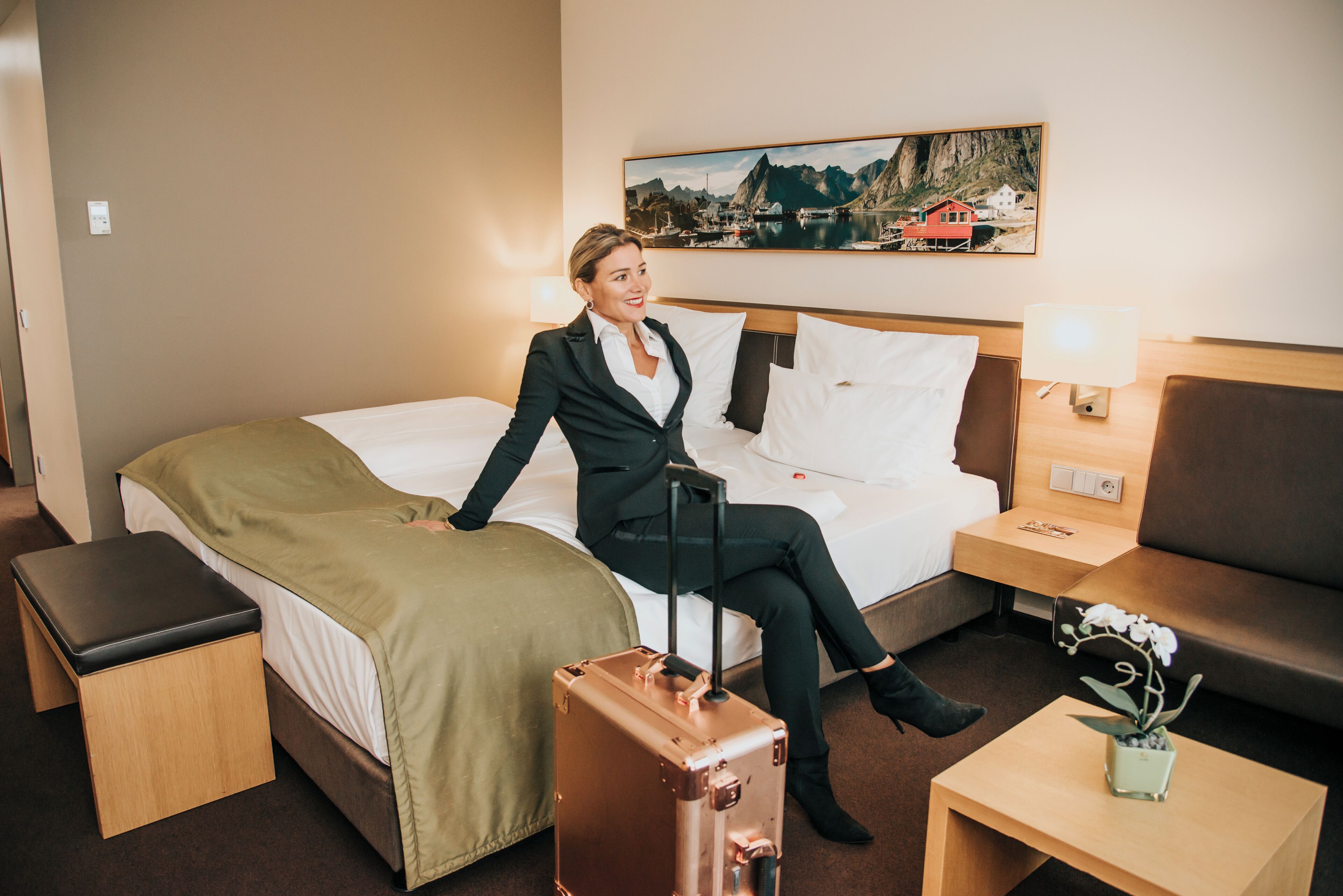 Ein Hotelzimmer mit einem Bett, einer hölzernen Bank, einem kleinen Tisch mit einer Lampe und einem Koffer.
