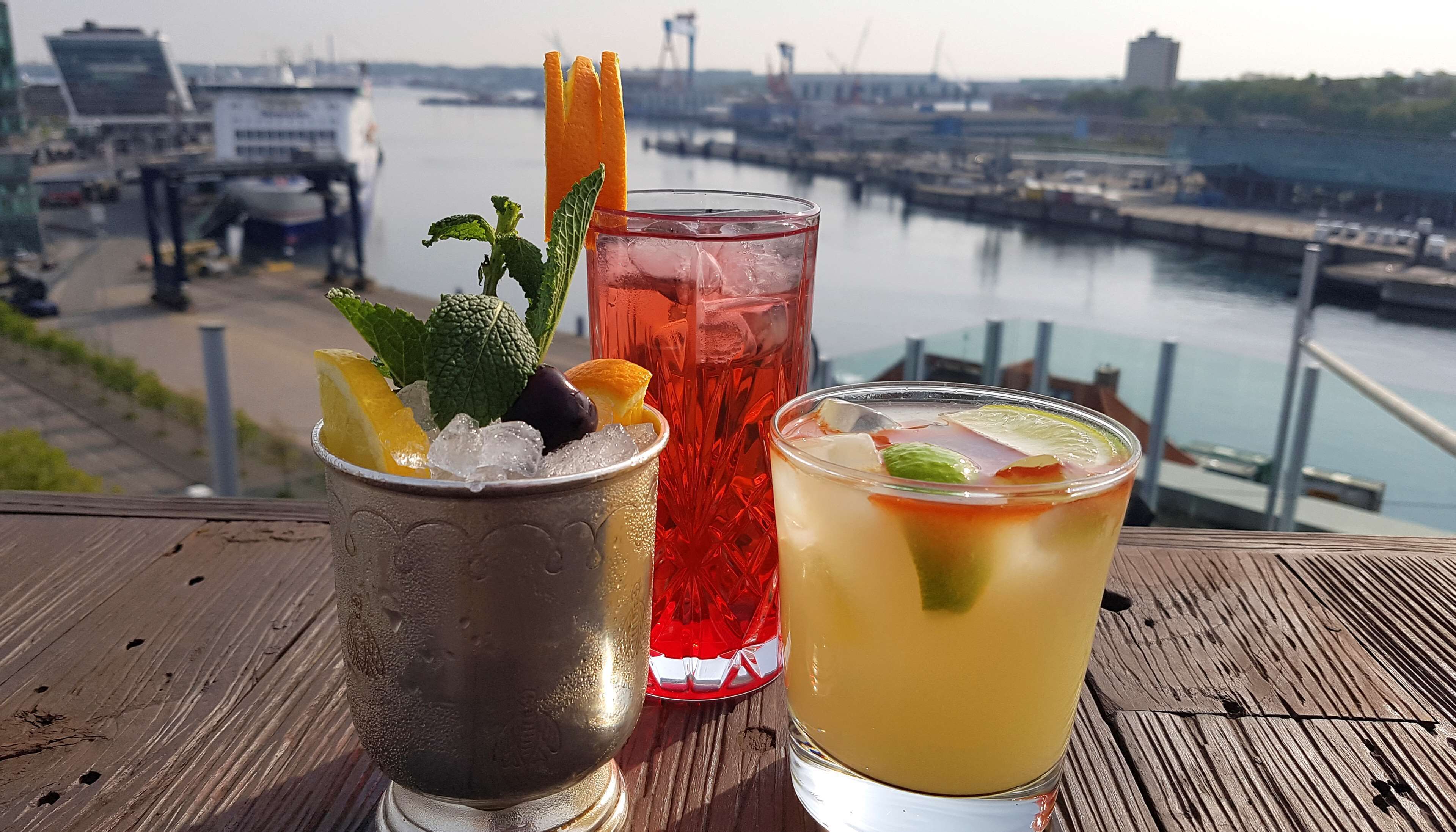 Three drinks on a wooden table with a view of a harbor and boats in the background.