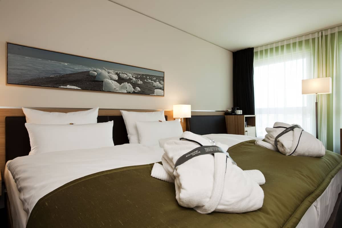 A hotel room with a bed, pillows, a blanket, a painting of a beach scene, a lamp, and a window with curtains.