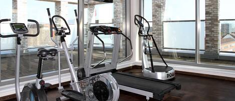 Un gimnasio con diferentes máquinas de ejercicio, una pantalla de televisión y amplios ventanales con vistas a edificios.