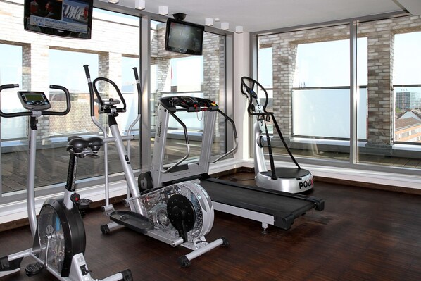 Un gimnasio con diferentes máquinas de ejercicio, una pantalla de televisión y amplios ventanales con vistas a edificios.