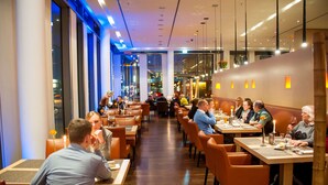 A modern restaurant with wooden tables and orange chairs, large windows, and a view of the city at night. - Atlantic Hotel Kiel (Kiel)