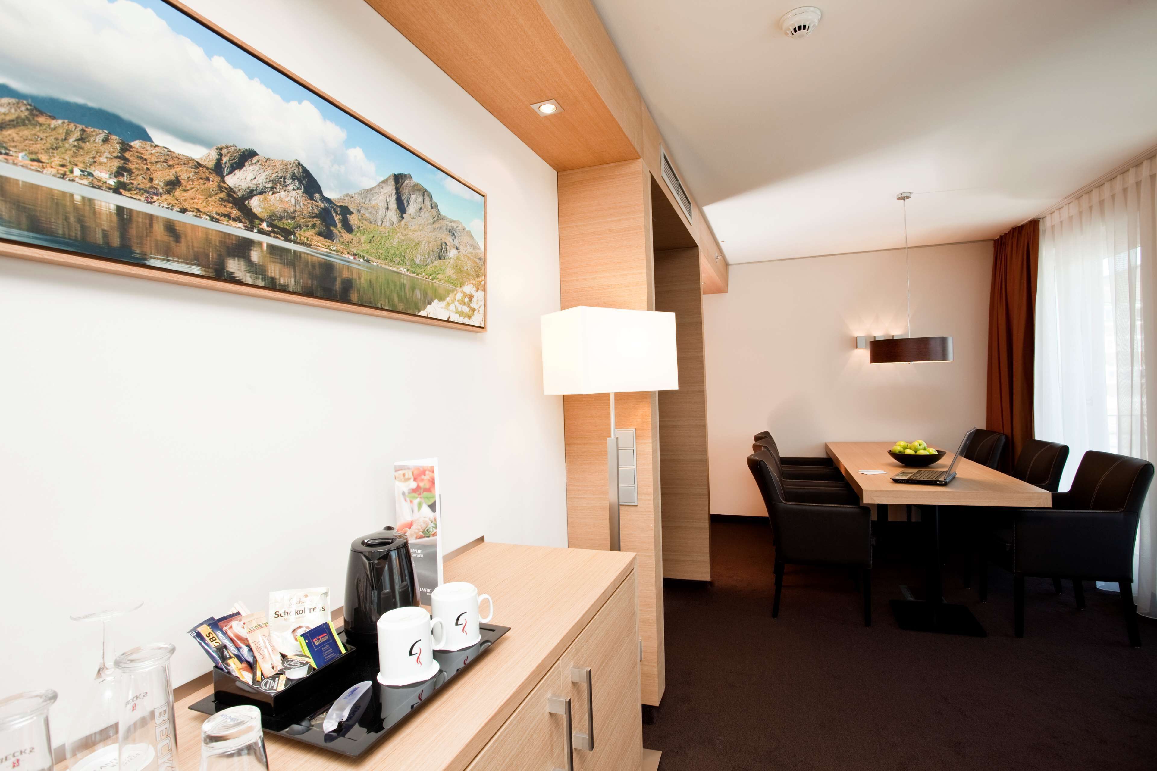 A hotel room with a wooden desk, a coffee machine, two cups, a tray, a framed picture of a mountain landscape, a dining table with chairs, and a lamp.