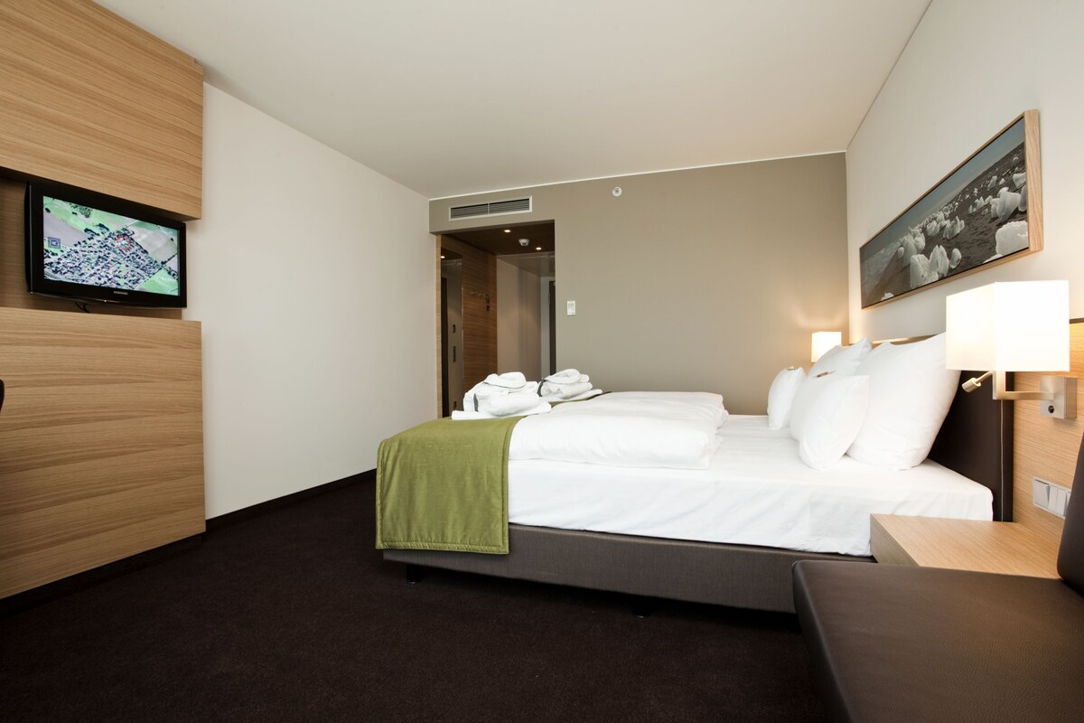 Una habitación de hotel con una cama, televisión, un cuadro y un escritorio con lámpara.