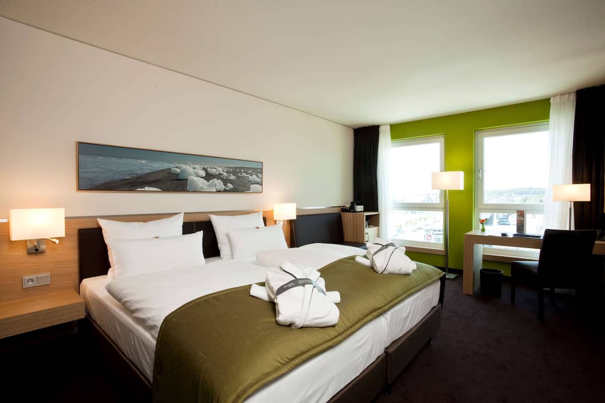 A hotel room with a large bed, two bedside tables with lamps, a desk with a chair, and a window with curtains.