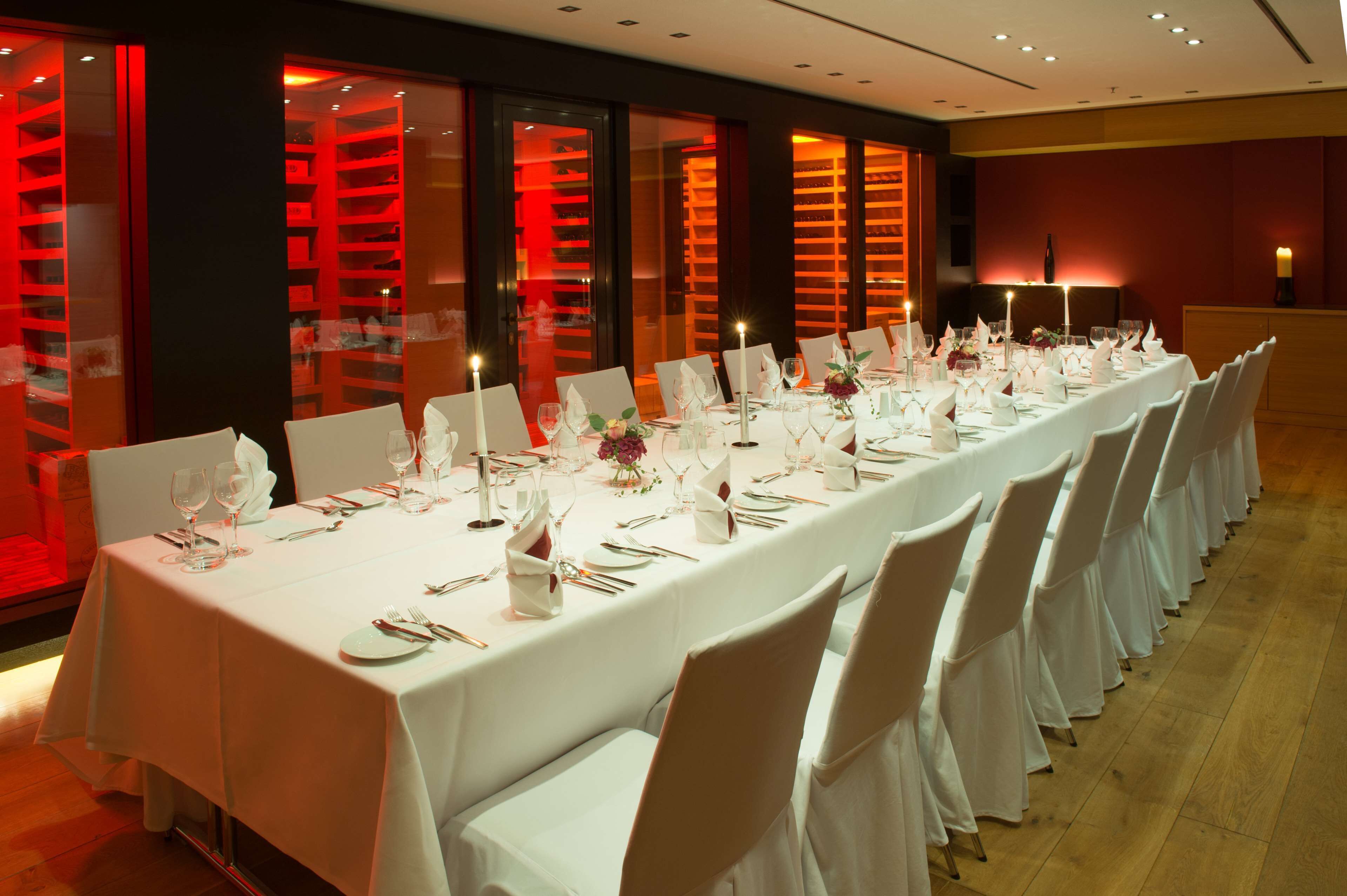 A long dining table set with white tablecloths, chairs, and candles, in a room with red-lit shelving units.