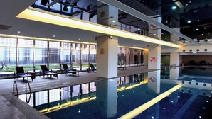 A modern indoor swimming pool with clear glass walls, lounge chairs, and a view of the outside.