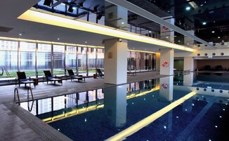 Una piscina interior moderna con paredes de vidrio transparente, sillas de descanso y vistas al exterior.