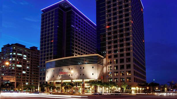 A hotel with a modern design, featuring a prominent 'Sheraton' sign. It has multiple floors with a grid-like facade and is surrounded by other high-rise buildings.