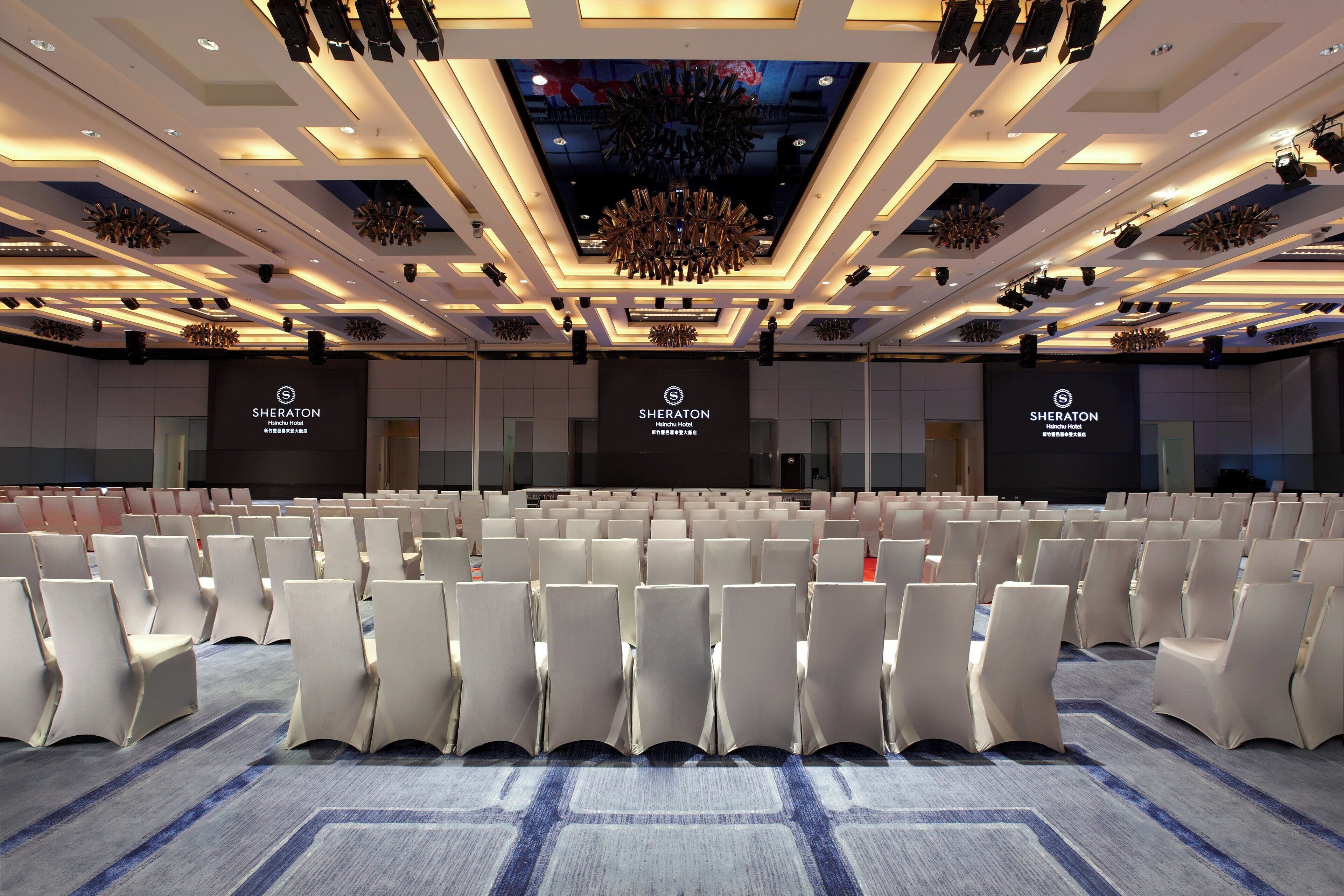 Une salle de conférence d’un hôtel Sheraton, avec des rangées de chaises blanches, une moquette à motifs et un design de plafond moderne.
