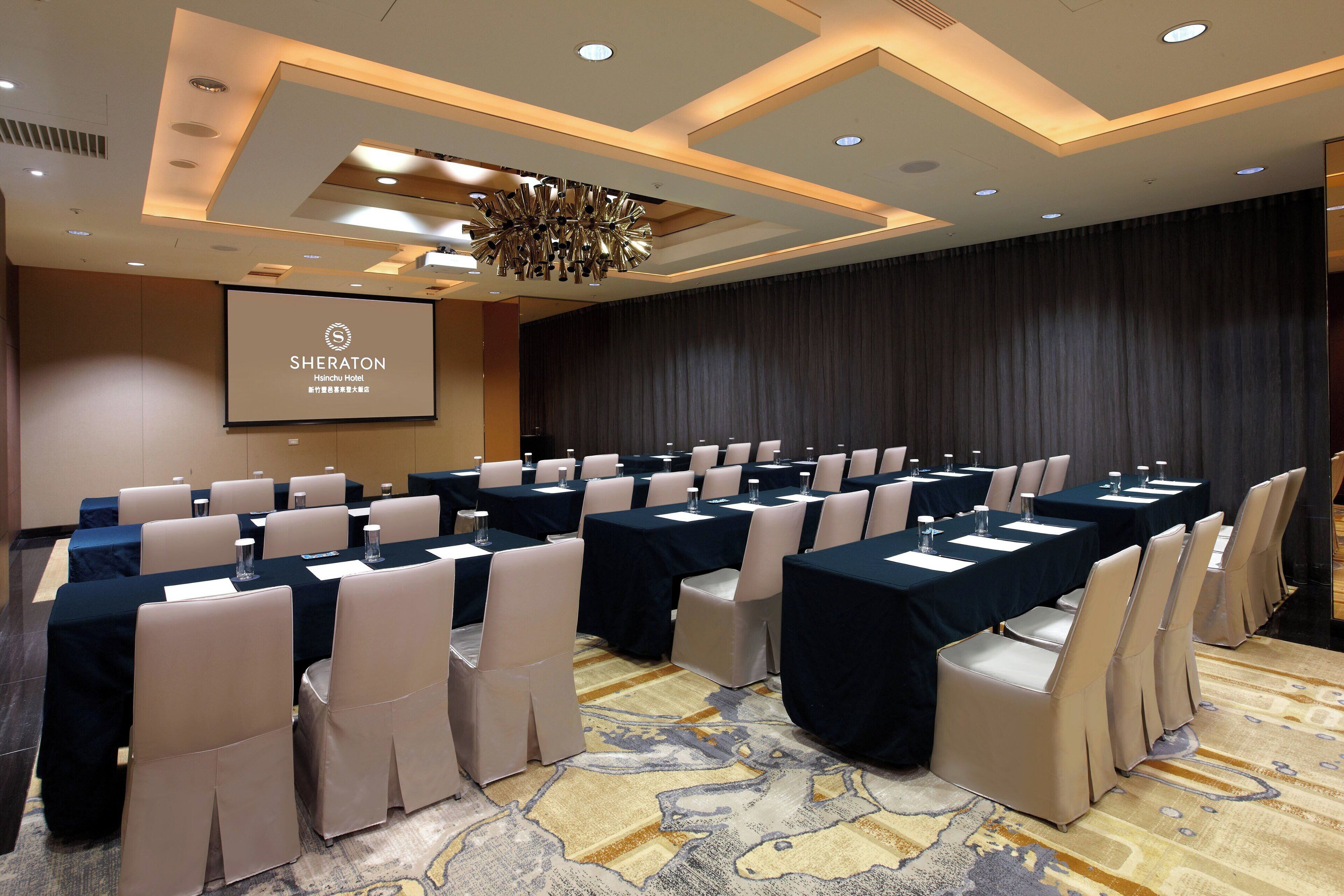 A conference room with rows of tables and chairs, a projector screen, and a chandelier.