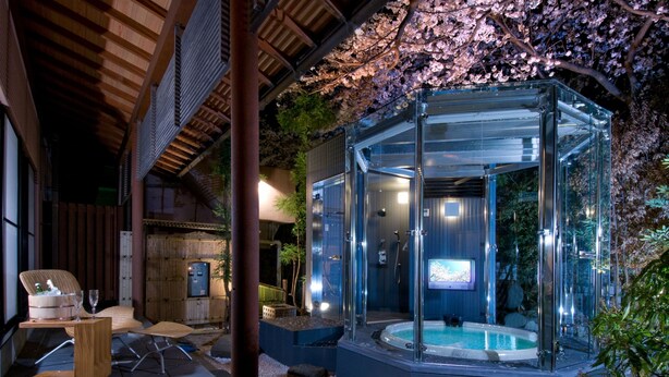 A glass-enclosed hot tub area with a wooden deck and seating, surrounded by cherry blossoms. - Shoheiso Ryokan (Izunokuni)
