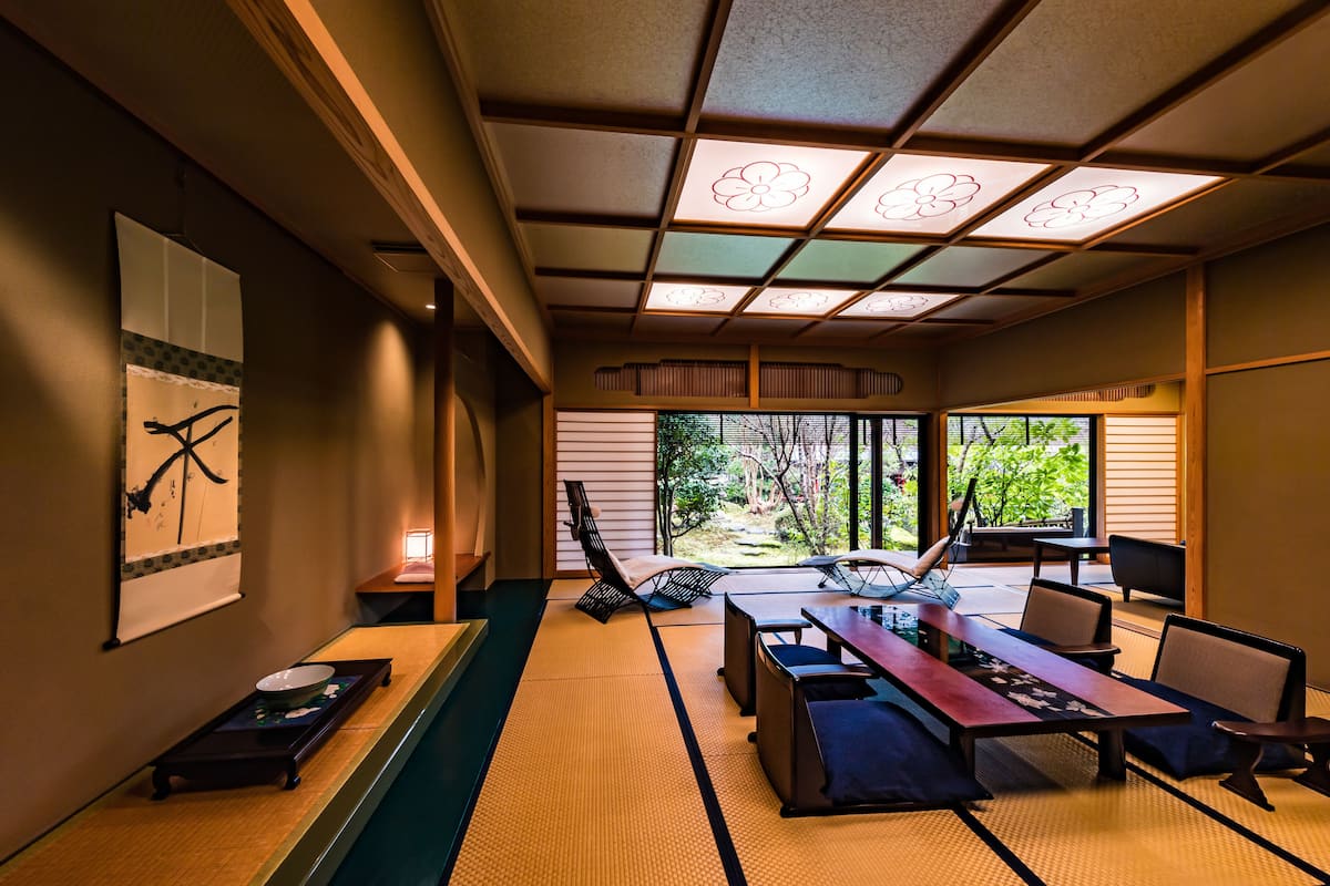 Una habitación japonesa tradicional con suelo de tatami, una mesa baja con cojines y vistas a un jardín a través de amplias ventanas.