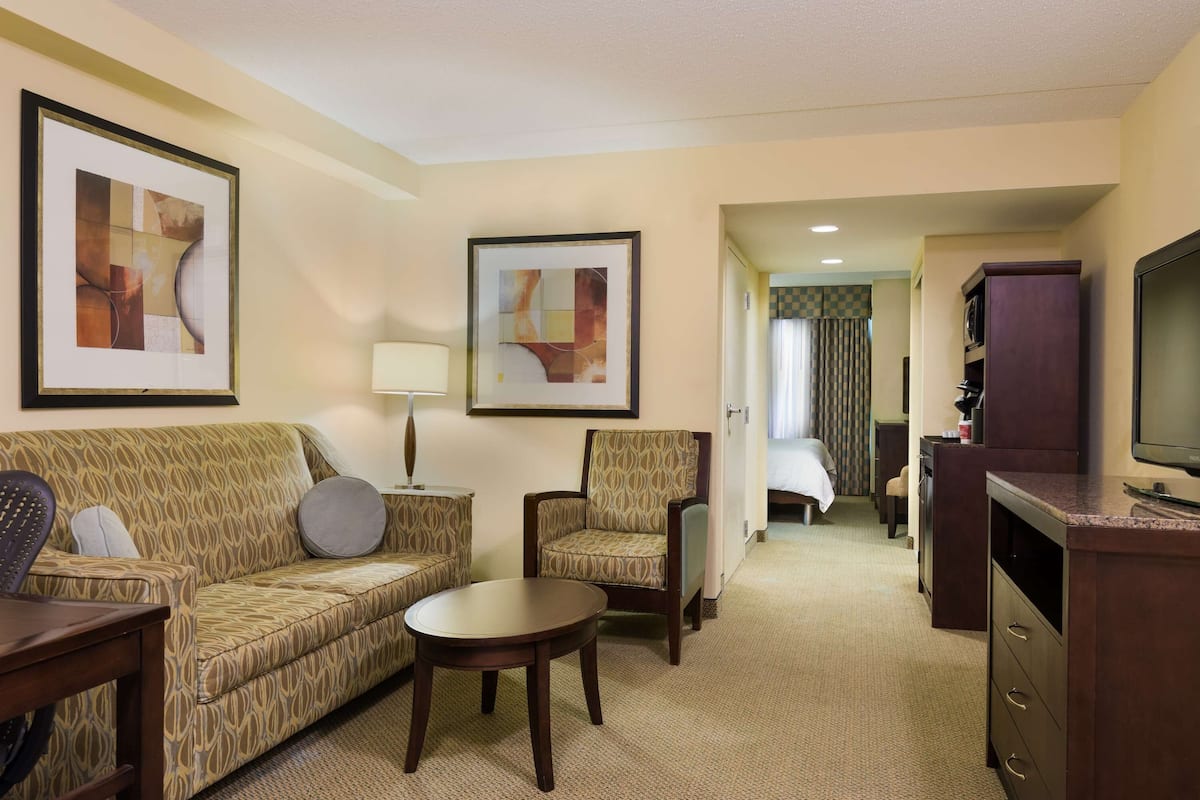 A hotel room with a patterned sofa, armchair, and a small round table. There is a television on a cabinet and framed artwork on the walls.