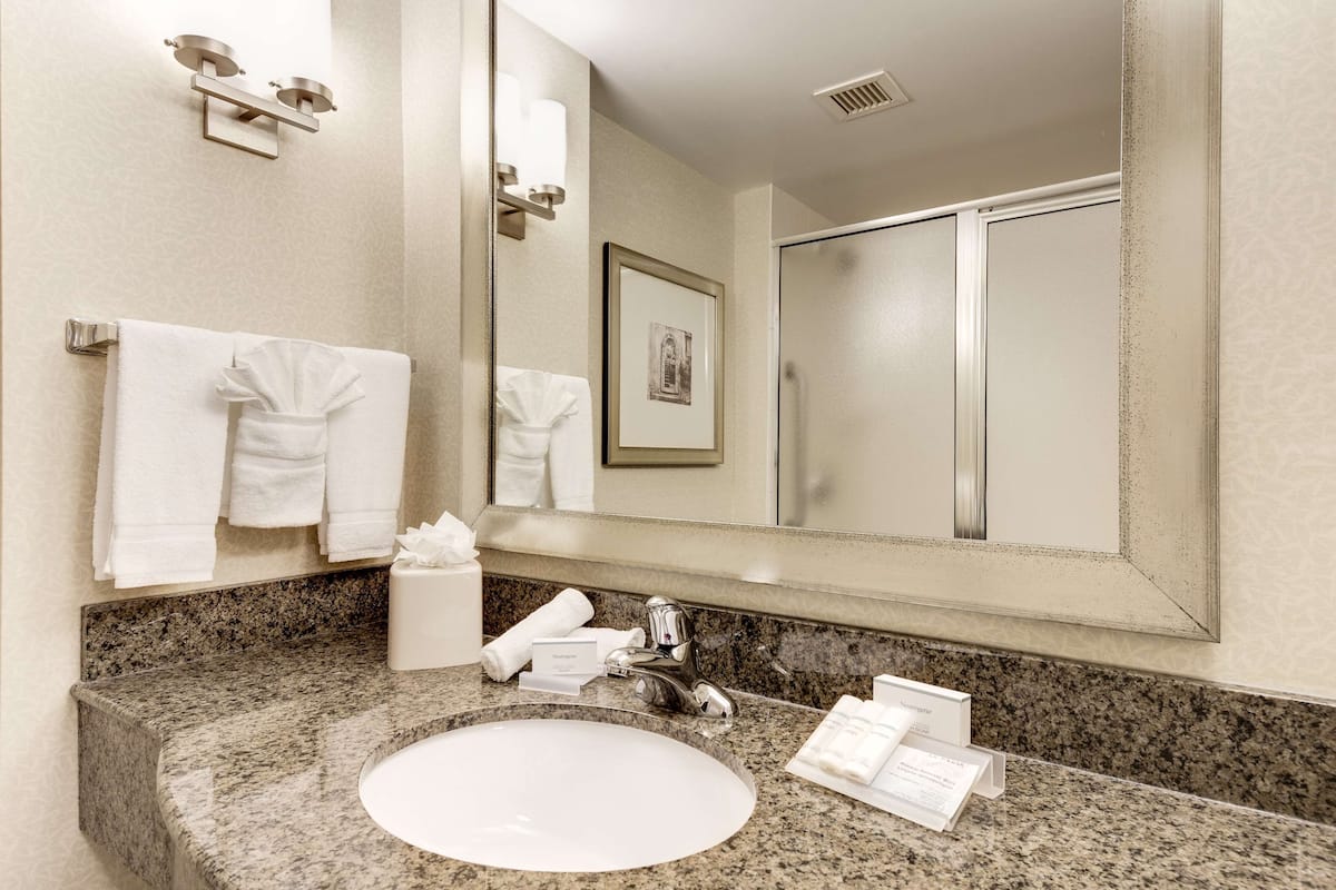 A hotel bathroom with a granite countertop, a white sink, a large mirror, and a framed picture on the wall.