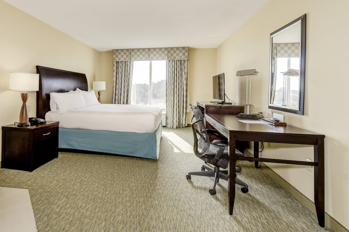 A hotel room with a bed, desk, chair, and a large window with curtains.