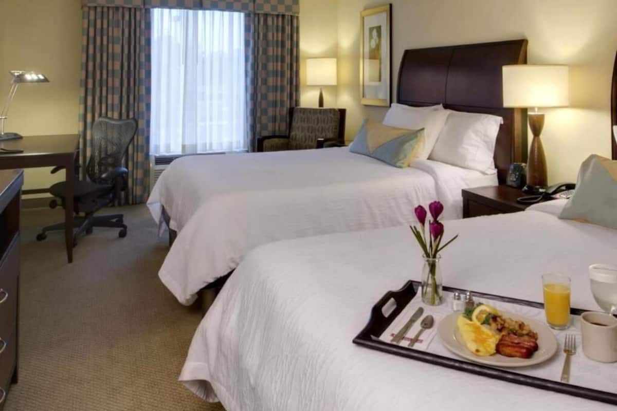 A hotel room with two beds, a desk, a chair, a dining table with breakfast, and a window with curtains.