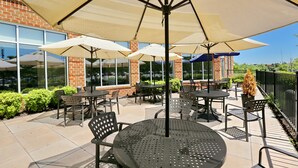 An outdoor patio with tables and chairs under umbrellas, a brick building in the background, and a black metal fence. - Hilton Garden Inn Waldorf (Waldorf)
