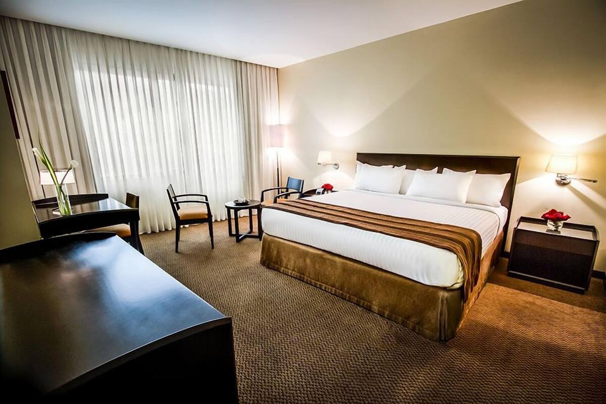 A hotel room with a large bed, a desk, a chair, a small table, and a nightstand with a lamp.