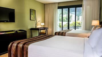 Habitación de hotel con dos camas, un escritorio, una silla, un televisor y una ventana con vistas a edificios y zonas verdes.