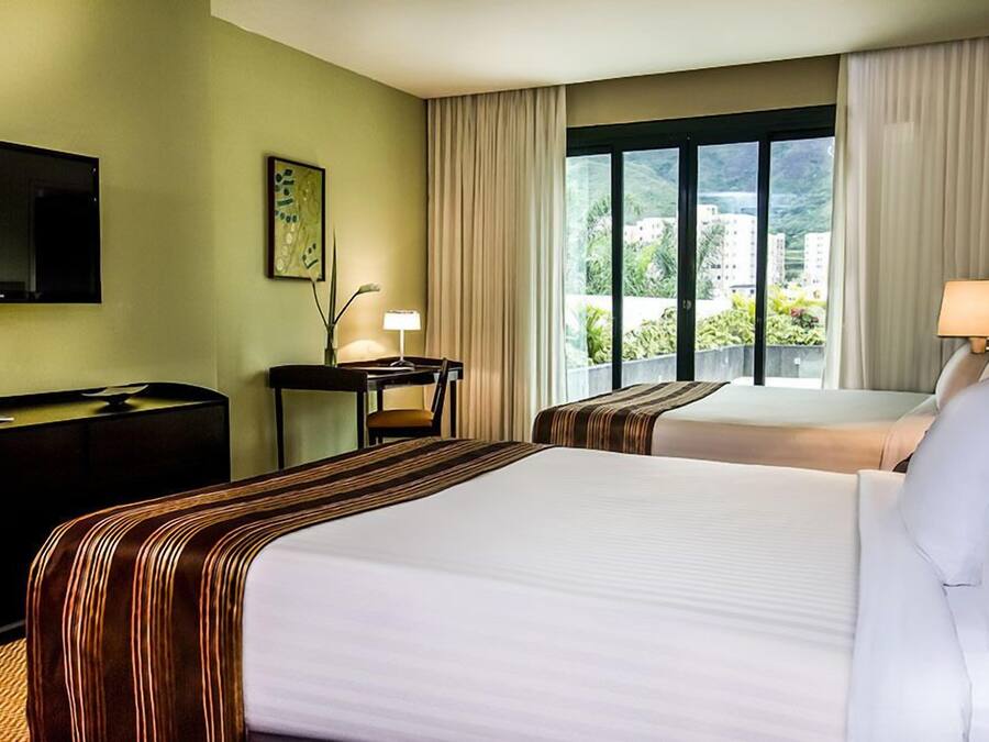 Habitación de hotel con dos camas, un escritorio, una silla, un televisor y una ventana con vistas a edificios y zonas verdes.