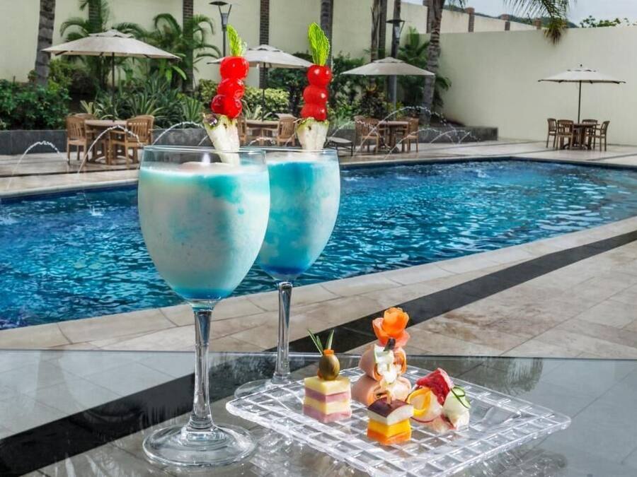 A poolside setting with two large glasses of blue cocktail, a tray of assorted desserts, and outdoor furniture.