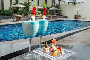 Ambiente junto à piscina com dois copos grandes de cocktail azul, um tabuleiro com sobremesas variadas e mobiliário exterior.