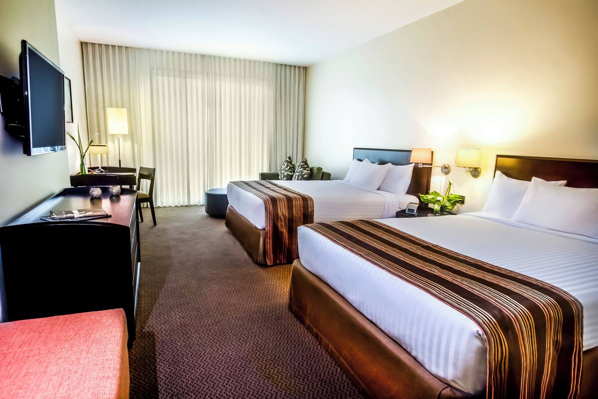 A hotel room with two beds, a desk, a TV, and a large window with curtains.