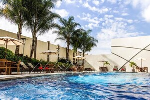 A swimming pool with lounge chairs and umbrellas, surrounded by palm trees and a white wall.