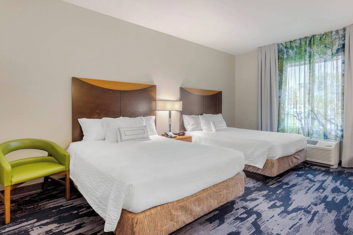 A hotel room with two beds, a green chair, a bedside table with a lamp, and a large window with curtains.