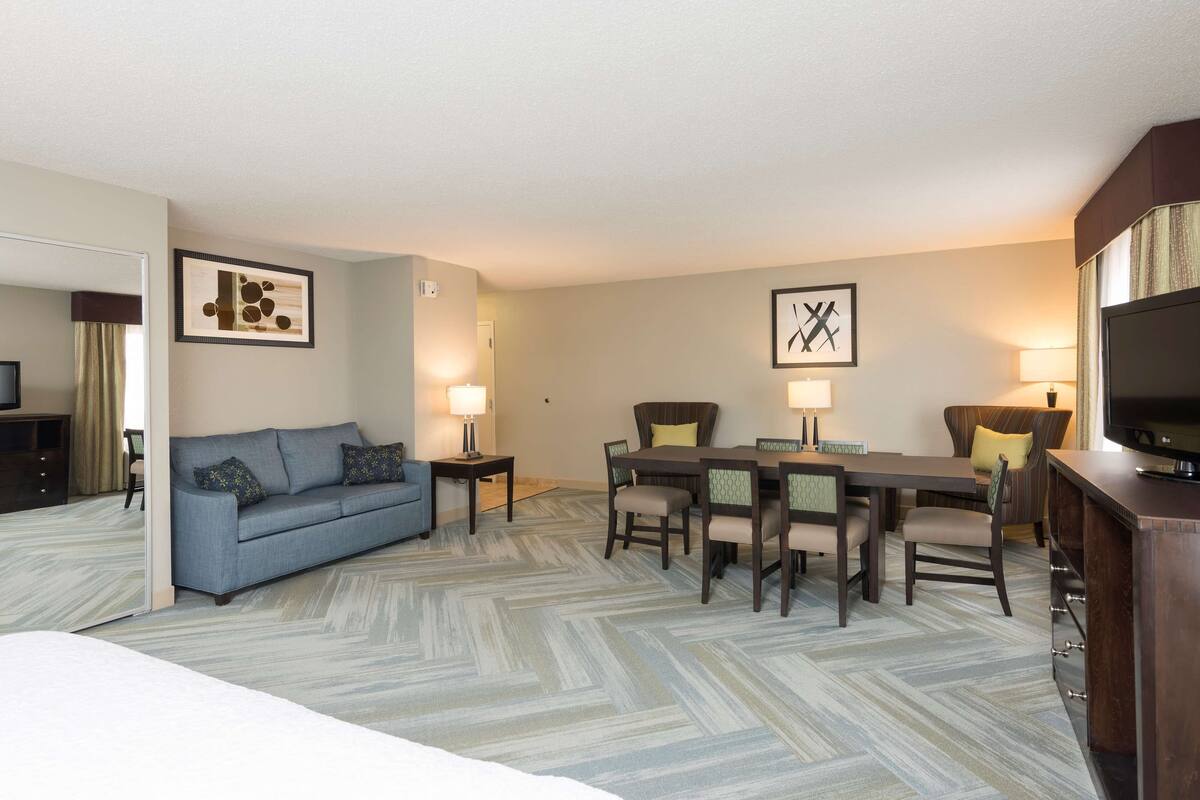 A hotel room with a bed, a sofa, a dining table with chairs, a TV, and artwork on the walls.