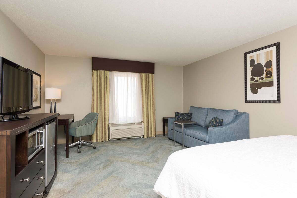 A hotel room with a bed, a TV, a desk with a lamp, a chair, a sofa, and a window with curtains.