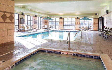 An indoor swimming pool with a hot tub, sunlit by large windows, surrounded by lounge chairs and umbrellas.