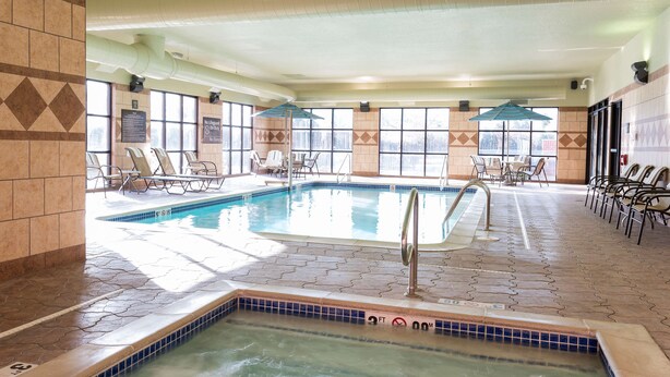 An indoor swimming pool with a hot tub, sunlit by large windows, surrounded by lounge chairs and umbrellas. - Hampton Inn Detroit/Roseville (Clinton Township)