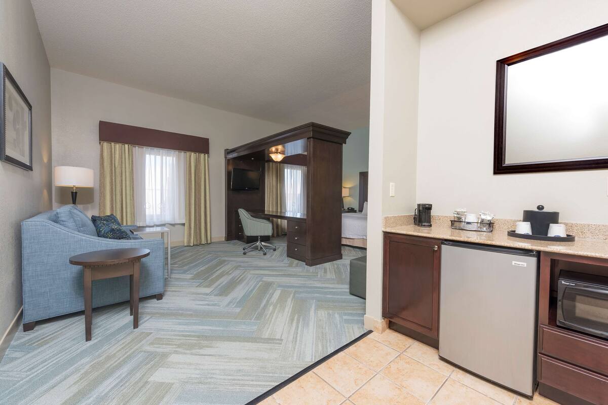 A hotel room with a sofa, a small table, a desk with a chair, a television, a kitchen area with a refrigerator and a microwave, and a large mirror on the wall.