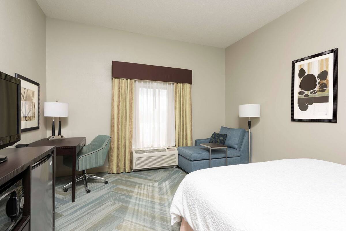 A hotel room with a bed, a desk with a lamp, a chair, a sofa, a TV, and a window with curtains.