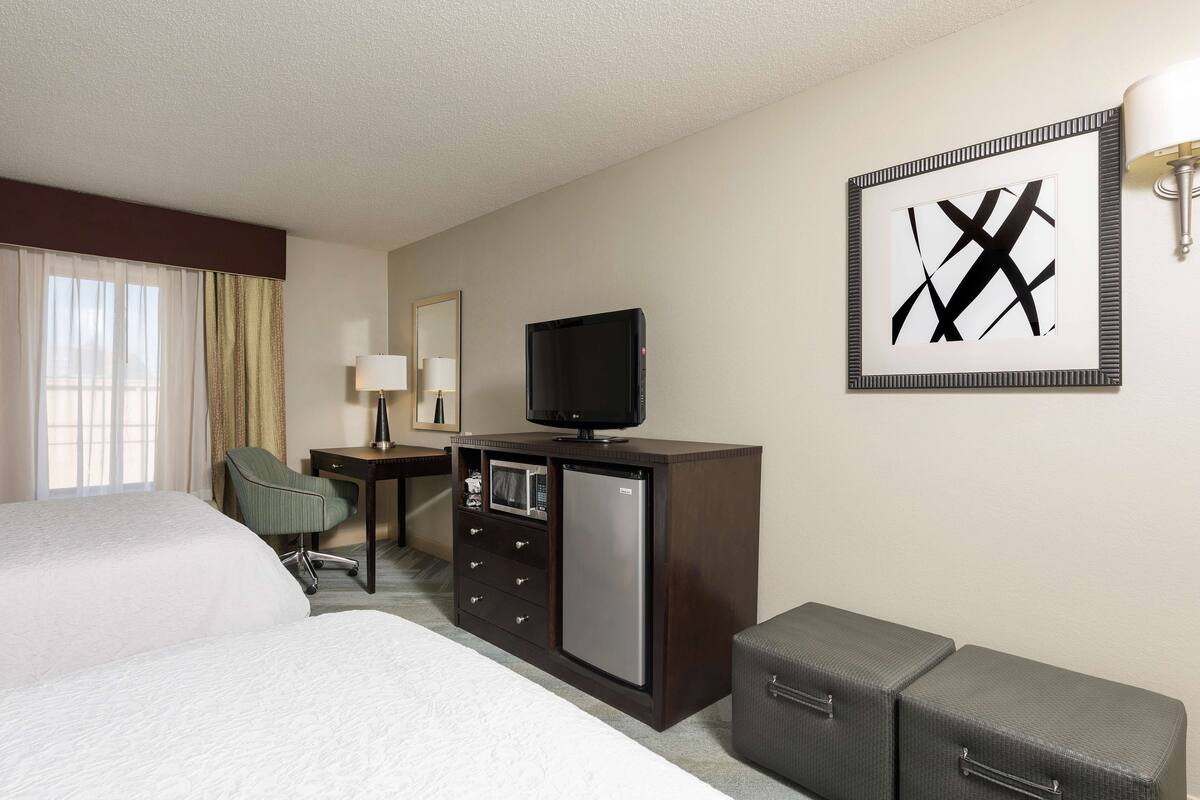 A hotel room with a bed, a desk with a television, a microwave, and a refrigerator. There is a chair, a lamp, and a framed artwork on the wall.