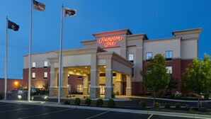 A Hampton Inn hotel with a prominent neon sign, multiple flags, and a well-lit entrance. - Hampton Inn Detroit/Roseville (Clinton Township)