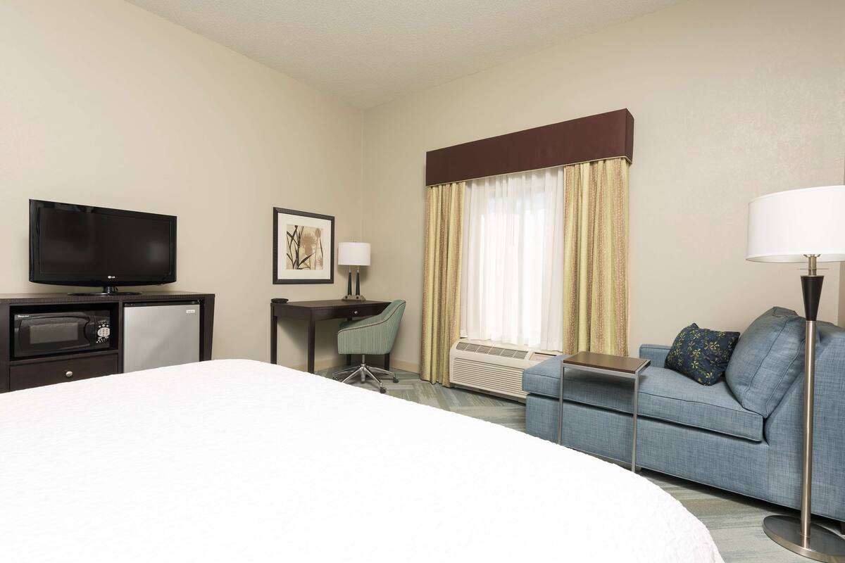 A hotel room with a bed, a desk with a computer, a television, a sofa, and a window with curtains.